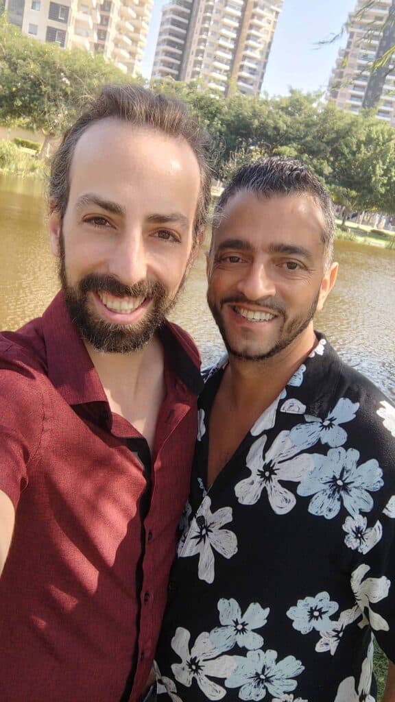 שי ונועם מחייכים יחד בפארק עם רקע של מים ועצים, ביום שמשי.